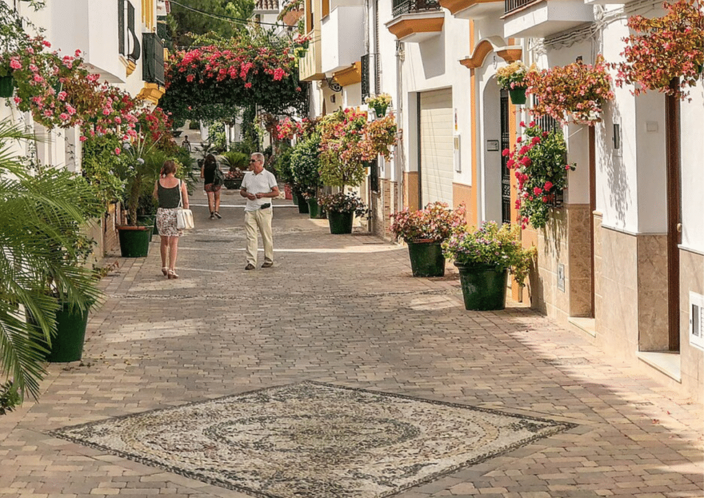 Flower‑lined cobblestone street with white buildings and yellow trim in a Spanish town.