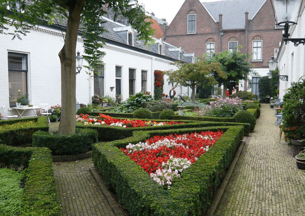 A hofje courtyard garden with shade trees, colorful flower beds, clipped hedges, and outdoor seating surrounded by white brick buildings.